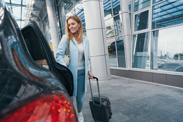 Smiling pleased woman with trolley suitcase closing door of taxicab parked before airport terminal building